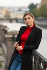 A portrait of young woman with brown long hear, in black coat and red top, blue jeans, standing by the fence on the embankment with cityscape and river on background / 