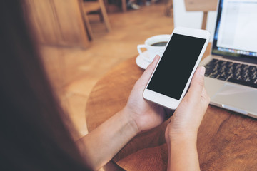 Mockup image of hands holding white smartphone with blank black screen ,laptop and coffee cup on wooden table in cafe