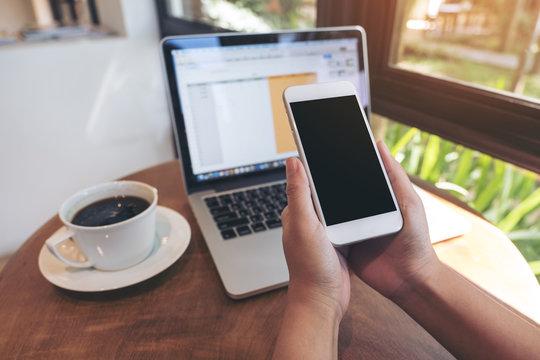 Mockup image of hands holding white smartphone with blank black screen ,laptop and coffee cup on wooden table in cafe