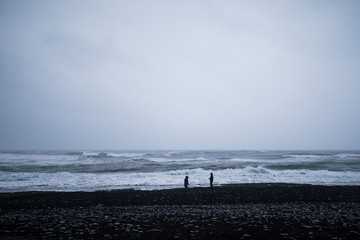 Two figures stand on a black sand beach in stormy weather. Iceland, Europe.