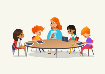 Redhead female teacher showing picture to children sitting around round table at class with laptop and tablet pc. Kids using gadgets during lesson at primary school. Colorful vector illustration.