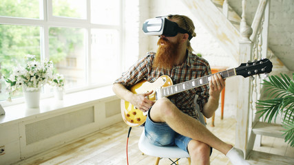 Young bearded man sitting on chair learning to play guitar using VR 360 headset and feels him...