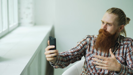 Bearded young hipster businessman talking video chat on smartphone while sitting in offce chair