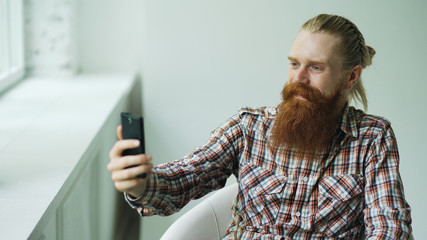Bearded young hipster businessman talking video chat on smartphone while sitting in offce chair