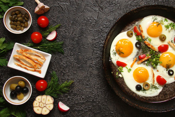 Fried eggs in a cast iron pan with peppers, tomatoes, capers, herbs and bread on a black background