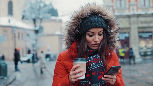 Beautiful happy woman walks around the city drinking coffee using the phone in winter smiling technology text weather white attractive brunette communication portrait close up snow slow motion