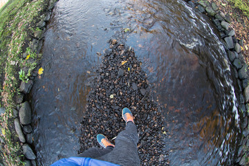 Man feet in sneakers standing on black stones in the middle of small river in the city park. Top view