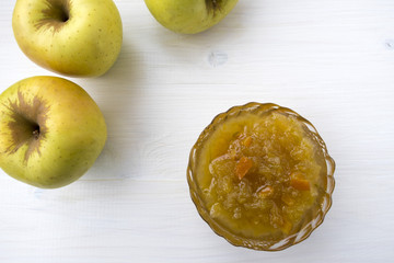 Apple jam and apples are located on a light wooden table.