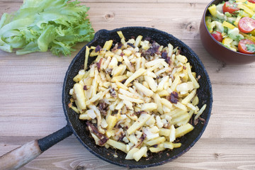 Fried potatoes in a rustic frying pan on a wooden background and vegetable salad.
