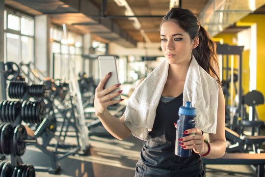 Sporty young woman with water bottle and towel around her neck using phone in gym.