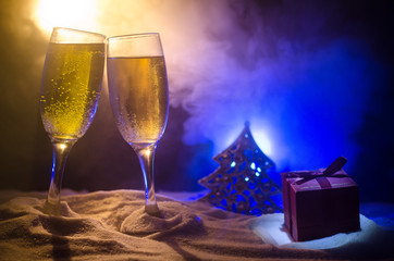 New Year Eve celebration background with pair of flutes and bottle of champagne with Christmas attributes (or elements) on snowy dark toned foggy background. Selective focus.