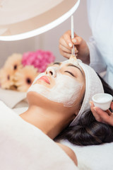 Close-up of the face of a young woman relaxing under the gentle touch of the specialist applying on her cheeks, white facial mask with rejuvenating effects