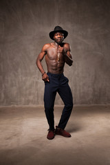 Fit strong physique African American young man wearing black  hat and  trousers posing dancing near gray concrete cement wall in studio.