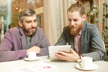 Portrait of two modern business people enjoying coffee break in cafe, using digital tablet via wireless connection and discussing work