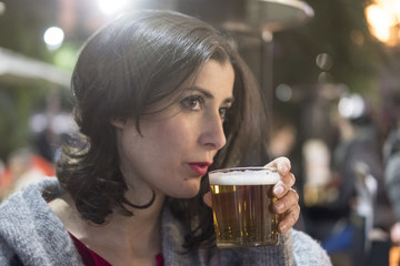 Beauty woman drinking cold beer in winter night with near a street stove of a bar terrace. Focus in beer glass