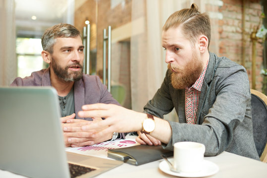 Portrait of two successful business men discussing work and using laptop during meeting in cafe on coffee break, focus on hipster businessman pointing to screen