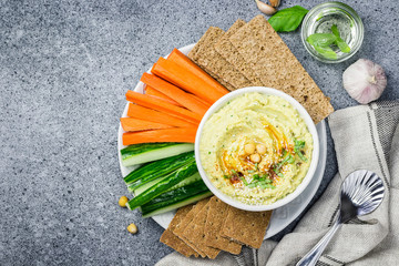 Classic smooth hummus in a bowl and carrot cucumber sticks on stone or concrete background. Top...