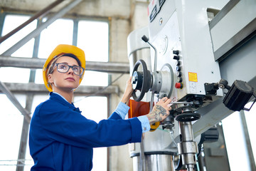 Portrait of attractive plant worker wearing eyeglasses and protective helmet operating bench drilling machine at spacious production department