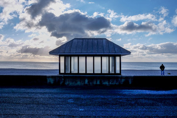 photograph of a modern glass and metal seating shelter on a pebble beach, sea behind and a cloudy contrasting sky, in the foreground are lines of tarmac. The image has a ‘graphical shapes’ look.
