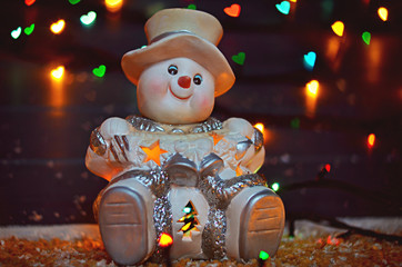 Ceramic snowman  candle holder  with garland lights on gray wooden background. Merry Christmas, heart bokeh, snow