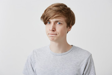 Casually dressed male model presses lips, looks pensively with blue eyes at camera, thinks over his future actions or plans, has confused expression, isolated against gray studio background
