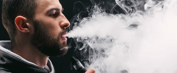 Young man vaping e-cigarette with smoke on black