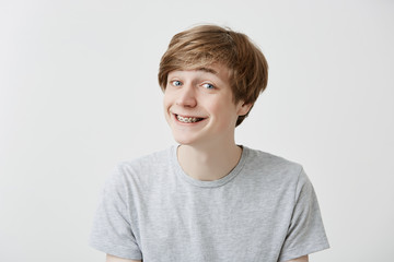 Cheerful caucasian guy grins at camera, has joyful expression, clenches teethwith braces, having fun with friends indoors. Smiling young male student in t-shirt expresses positiveness