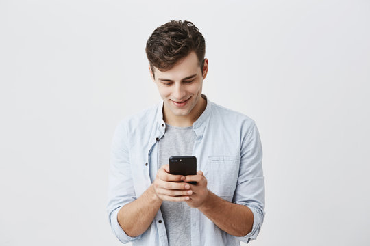 Handsome joyful student with dark hair wearing blue shirt messaging, typing message, using free onlipe app on his smartphone, looking at screen with smile, posing against gray background.