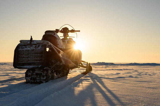 A Snowmobile Stands On The Snow At Sunset.