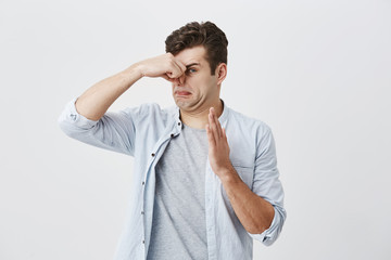 Caucasian male hipster with dark hair dressed in light blue shirt over gray t-shirt pinching nose...