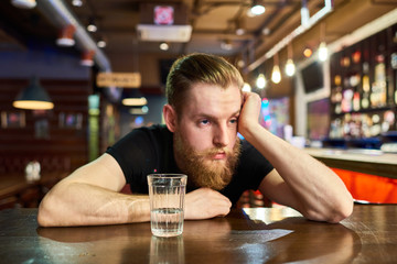 Front view portrait of drunk bearded man in pub sitting at table with glass of vodka looking away...