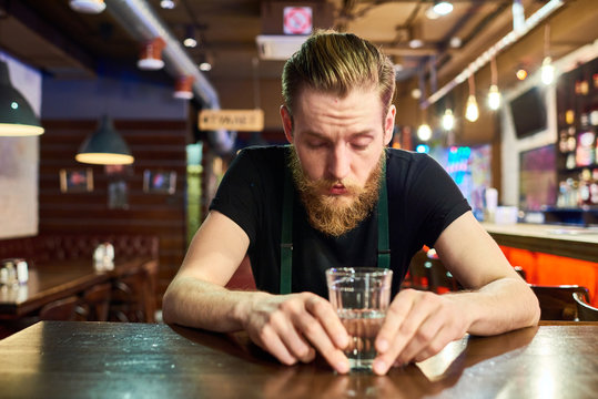 Front view portrait of modern bearded man getting drunk alone in pub sitting at table and drinking vodka