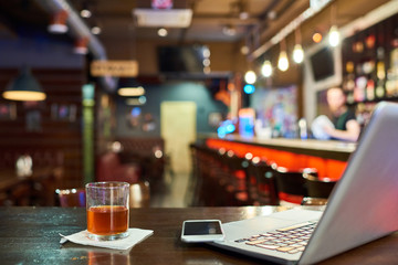 Background image of empty workplace in pub, laptop on bar counter next to glass of scotch or whiskey, remote and freelance work concept