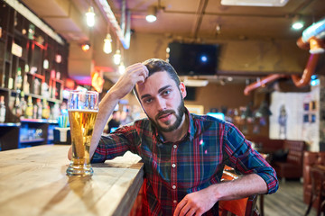 Portrait of modern  bearded  man sitting at bar counter with tall glass of beer and looking at camera, copy space