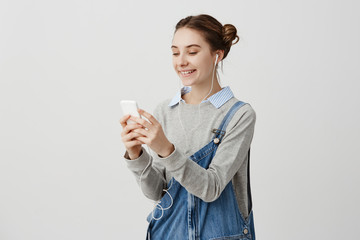 Happy girl wearing denim overalls standing in earphones outside having Skype call. Good-looking...