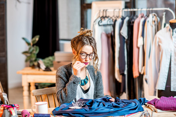 Young female fashion designer working with blue fabric sitting at the beautiful office with different tailoring tools on the table