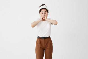 Indoor shot of scared dark-haired female with hairbun looks with bugged eyes and opened mouth, frightened to see awful things, can`t believe her eyes. People, emotions, feelings concept