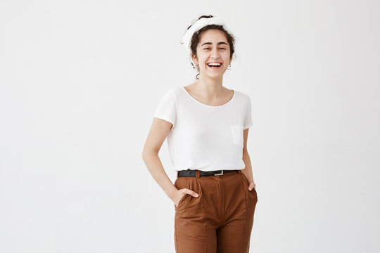 Pleased pretty woman with dark and wavy hair in bun, dark eyes and healthy skin dressed in white T-shirt and brown trousers holding hands in pockets, posing against white background.