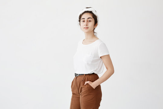 Attractive young female model with dark and wavy hair in bun, wearing white t-shirt and trousers, keeping her hands in pockets, posing against white background with copy space for advertisment