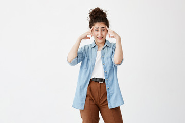 Brunette female with hair bun has headache after noisy party, clenches teeth and holds index fingers on temples. Tired young female worker overworked after writing advertising content