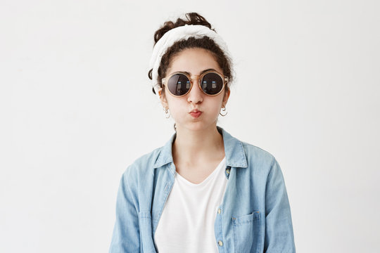 Funny female model with dark hair in bun pouts lips, in denim shirt and roung sunglasses, makes grimace as stands in studio. Joyful pretty woman has fun indoors, blows cheeks, foolishes at camera