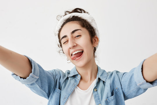 Happiness, beauty, joy and youth. Young positive girl dressed in denim shirt over white t-shirt stretching arms, smiling broadly, blinking, sticking out her tongue, having good mood.