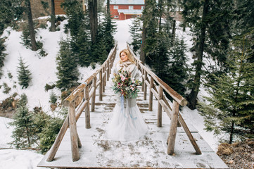 Beautiful woman stands on the wooden bridge in snowy forest in grey wedding dress with bouquet in...