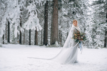 Beautiful woman stands on the snowy pine forest in grey wedding dress with bouquet in hands and long veil
