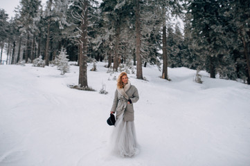 Beautiful blonde woman in hat and grey wedding dress and coat is walking on the winter landscape
