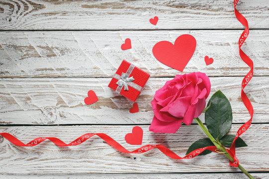 Rose with red paper hearts and gift box and ribbon on white wooden background. Love symbol concept.