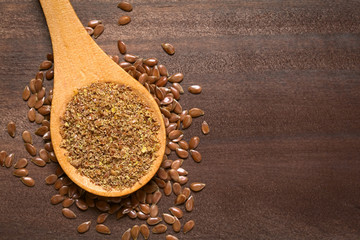 Ground or crushed brown flax seed or linseed on wooden spoon, photographed on dark wood with natural light - obrazy, fototapety, plakaty