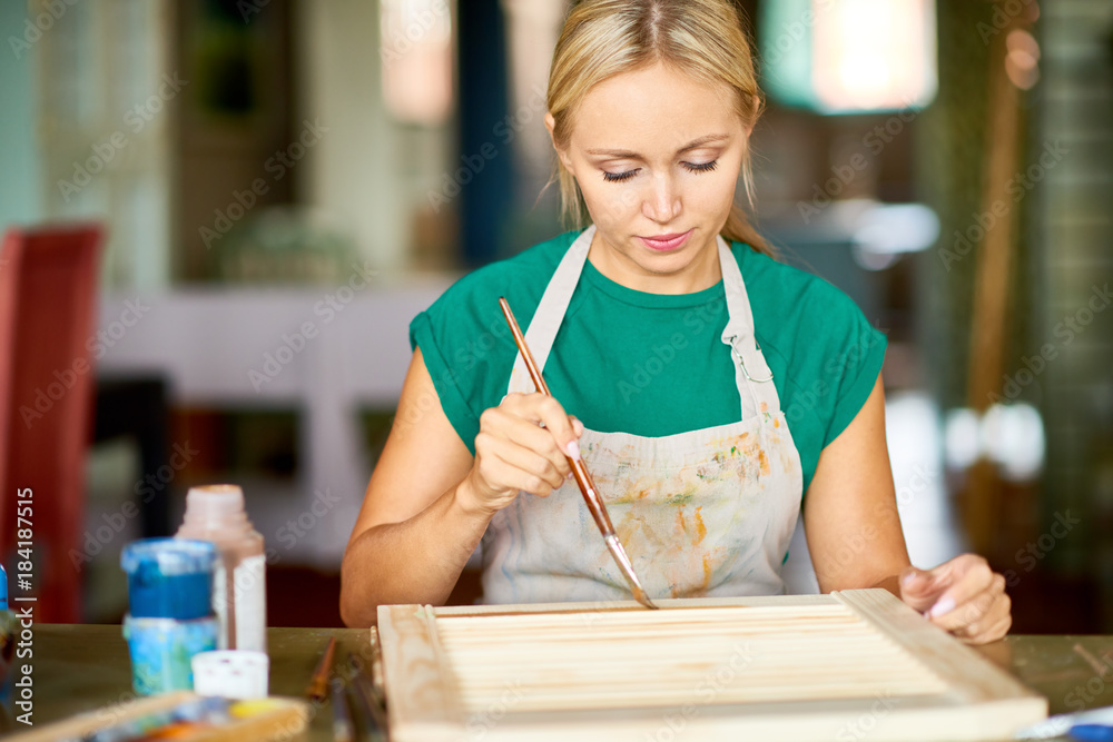 Wall mural Front view portrait of pretty blonde woman enjoying work in art studio painting frame with silver paint, making DIY interior decoration