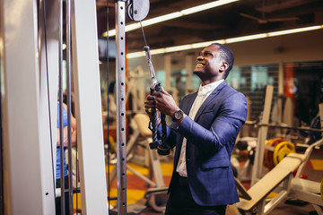 Muscular african man in classic suit working out in gym doing exercises at triceps, strong male businessman