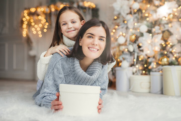Indoor shot of mother and daughter have fun together, share presents, being in room decorated with garlands and Christmas tree, have joyful expressions, enjoy weekends and winter holidays